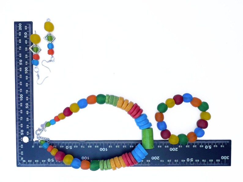 A vibrant jewellery set comprising a necklace, bracelet, and earrings. The necklace and bracelet feature round, multi-coloured beads. The earrings have yellow and green details. The jewellery is displayed on a white surface against a ruler to show the scale.