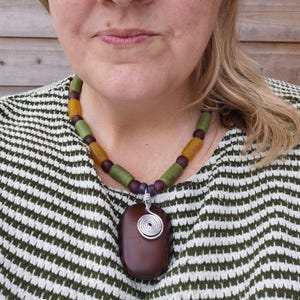 Peut inclure: Un collier fait &agrave; la main avec un pendentif ovale marron fonc&eacute; et une spirale en fil d'argent. Le collier est compos&eacute; de perles vertes, jaunes et violettes. La personne porte un pull ray&eacute; vert et blanc. Bijou unique.