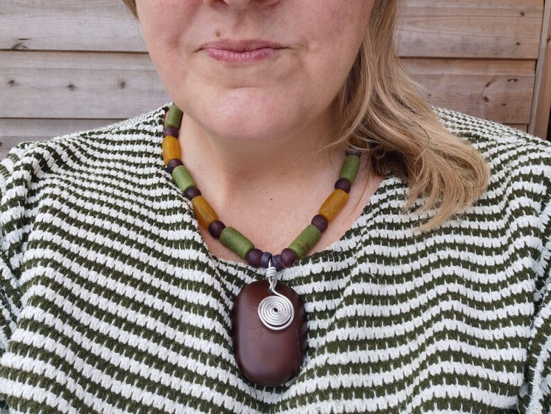 Peut inclure: Un collier fait &agrave; la main avec un pendentif ovale marron fonc&eacute; et une spirale en fil d'argent. Le collier est compos&eacute; de perles vertes, jaunes et violettes. La personne porte un pull ray&eacute; vert et blanc. Bijou unique.