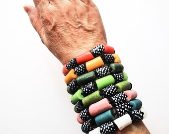 Puede incluir: Una pulsera de múltiples hilos con cuentas cilíndricas de varios colores sólidos, incluyendo rojo, naranja, verde, rosa, azul y blanco, que se alternan con cuentas negras más pequeñas decoradas con un patrón de lunares blancos.