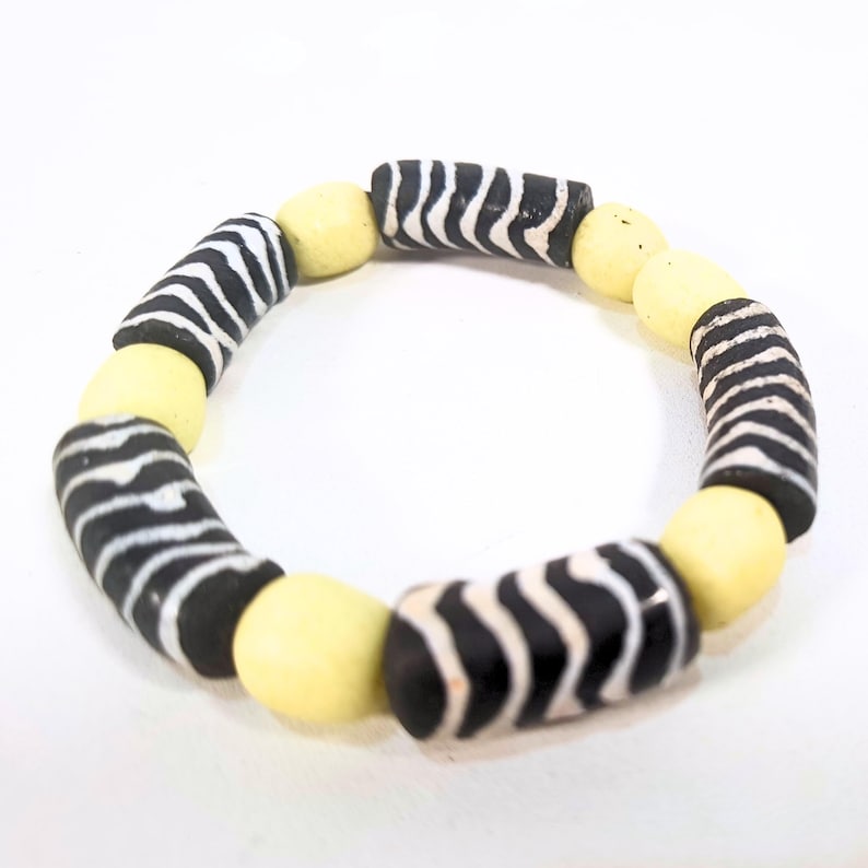 May include: A beaded bracelet featuring alternating black and white striped cylindrical beads and pale yellow, rounded beads. The bracelet is displayed on a white background, showcasing its contrasting colors and patterns.