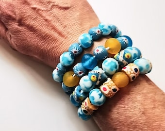 Puede incluir: Una pulsera de varias hebras con una variedad de cuentas redondas en tonos azul, blanco y amarillo. Las cuentas incluyen patrones marmoleados en azul y blanco, esferas amarillas mate sólidas y cuentas cilíndricas con diseños florales.