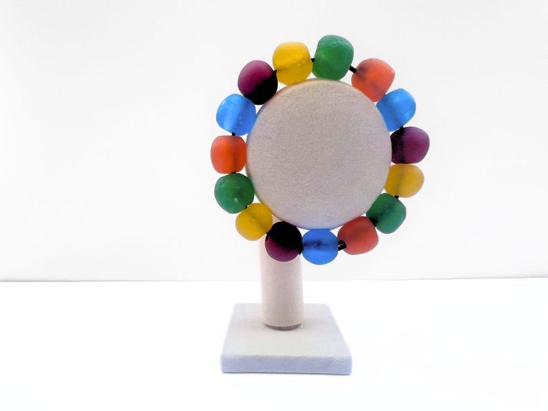 A vibrant beaded bracelet featuring a spectrum of recycled glass round beads, including red, orange, yellow, green, blue, and purple. The bracelet is displayed on a white, round jewellery stand with a square base.