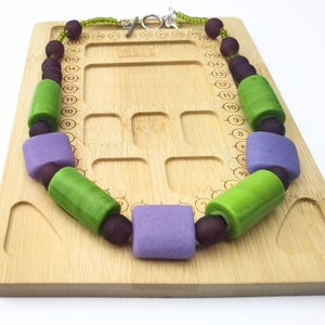 Puede incluir: Un collar colorido con grandes cuentas geom&eacute;tricas. El collar tiene cuentas verdes, moradas y rojo oscuro. El collar se exhibe sobre una superficie de madera marr&oacute;n claro. Cierre plateado.
