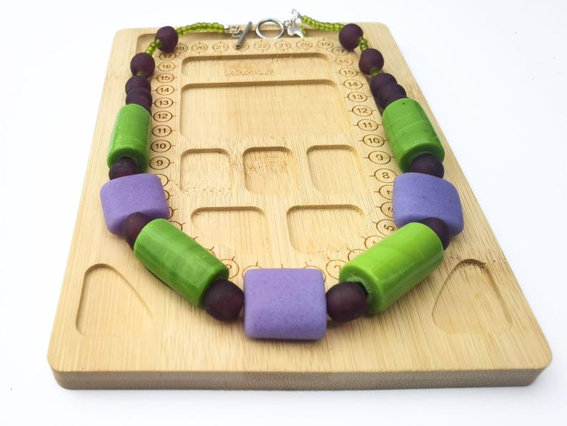 Puede incluir: Un collar colorido con grandes cuentas geom&eacute;tricas. El collar tiene cuentas verdes, moradas y rojo oscuro. El collar se exhibe sobre una superficie de madera marr&oacute;n claro. Cierre plateado.