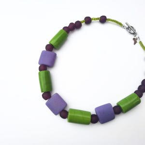 Puede incluir: Un collar con cuentas rectangulares verdes y moradas, intercaladas con cuentas moradas oscuras m&aacute;s peque&ntilde;as. El collar tiene un cierre plateado y un peque&ntilde;o dije. Las cuentas est&aacute;n ensartadas en un cord&oacute;n verde.