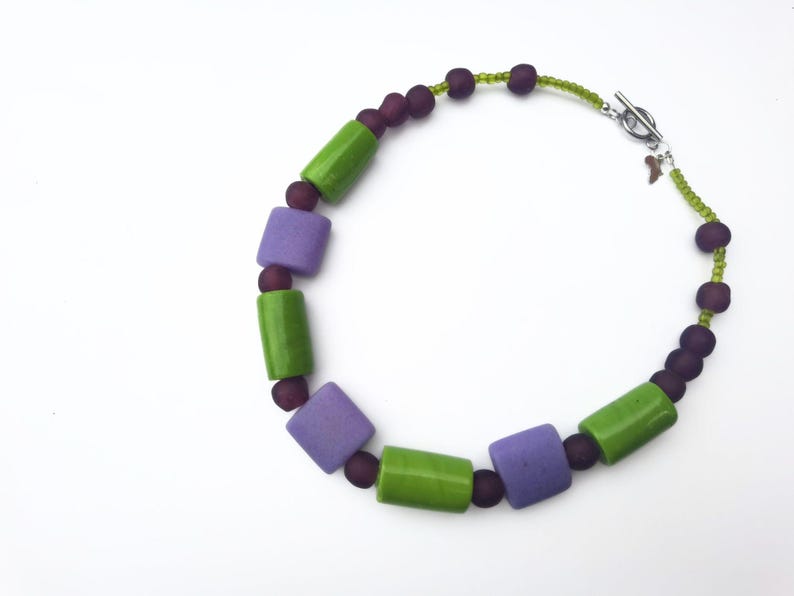 Puede incluir: Un collar con cuentas rectangulares verdes y moradas, intercaladas con cuentas moradas oscuras m&aacute;s peque&ntilde;as. El collar tiene un cierre plateado y un peque&ntilde;o dije. Las cuentas est&aacute;n ensartadas en un cord&oacute;n verde.