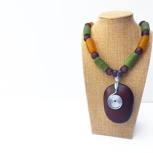 Peut inclure: Un collier artisanal avec un pendentif ovale marron fonc&eacute; orn&eacute; d'un motif en spirale en fil d'argent. Le collier est compos&eacute; d'une cha&icirc;ne de perles vertes, orange et violettes. Le collier est pr&eacute;sent&eacute; sur un pr&eacute;sentoir &agrave; bijoux en toile de jute.