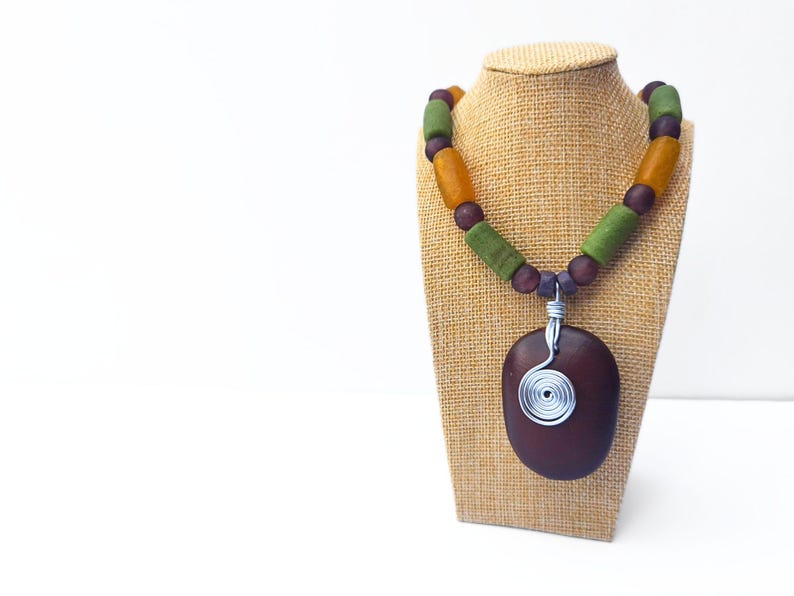 Peut inclure: Un collier artisanal avec un pendentif ovale marron fonc&eacute; orn&eacute; d'un motif en spirale en fil d'argent. Le collier est compos&eacute; d'une cha&icirc;ne de perles vertes, orange et violettes. Le collier est pr&eacute;sent&eacute; sur un pr&eacute;sentoir &agrave; bijoux en toile de jute.