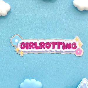 Könnte beinhalten: Aufkleber mit dem Wort "GIRLROTTING" in Pink, silber umrandet, mit hellblauem und rosa Blumenmuster auf hellblauem Hintergrund. Der Aufkleber hat einen weißen Rand und ist mit kleinen Blumen und Sternen verziert.