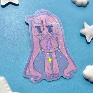 May include: A holographic sticker featuring a cartoon character with long pink hair, a knife in their head, and a blood stain. The character is wearing a pink outfit and has a star-shaped highlight. The sticker is surrounded by star and cloud-shaped decorations.