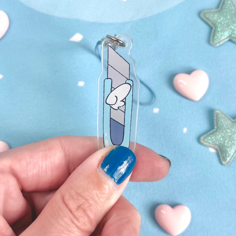 May include: A clear acrylic charm in the shape of a utility knife. The charm features a light blue and gray design with a white cartoon-style wing. A silver-colored ring is attached at the top for hanging.