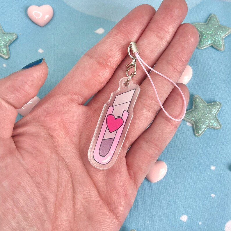 May include: A clear acrylic charm in the shape of a utility knife. The charm features a pink and purple color scheme with a red heart design. It includes a silver-colored clasp and a pink string for attachment.