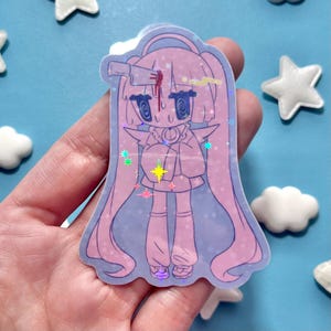 May include: A holographic sticker featuring a cartoon character with long pink hair and a knife in their head. The character has large eyes and is wearing a pink outfit. The sticker has a light blue background with star and cloud-shaped decorations.