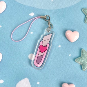 May include: A clear acrylic charm shaped like a utility knife, featuring a pink and purple design with a heart. It has a pink lanyard and a silver clasp. The charm is on a light blue background with heart and star-shaped decorations.