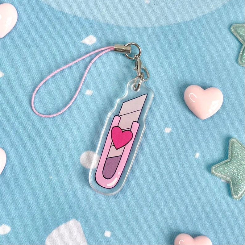 May include: A clear acrylic charm shaped like a utility knife, featuring a pink and purple design with a heart. It has a pink lanyard and a silver clasp. The charm is on a light blue background with heart and star-shaped decorations.