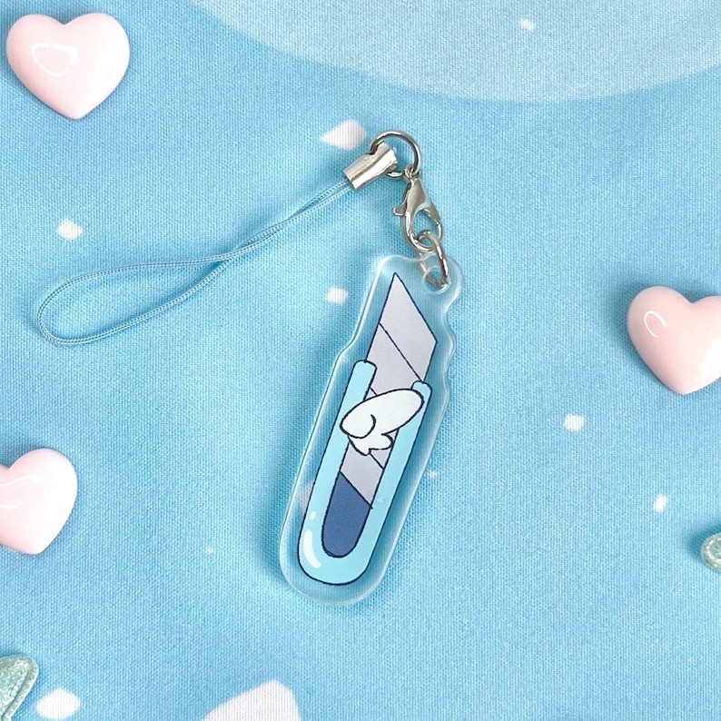 May include: A clear acrylic charm featuring a cartoon utility knife design with a light blue and gray color scheme. The charm has a silver-colored clasp and a matching wrist strap. The background is a light blue fabric with pink heart-shaped decorations.