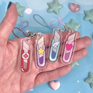 May include: Four clear acrylic charms shaped like utility knives, each with a different design: a red cross, a yellow star, a fish, and a pink heart. Each charm has a silver clasp and a colored string.