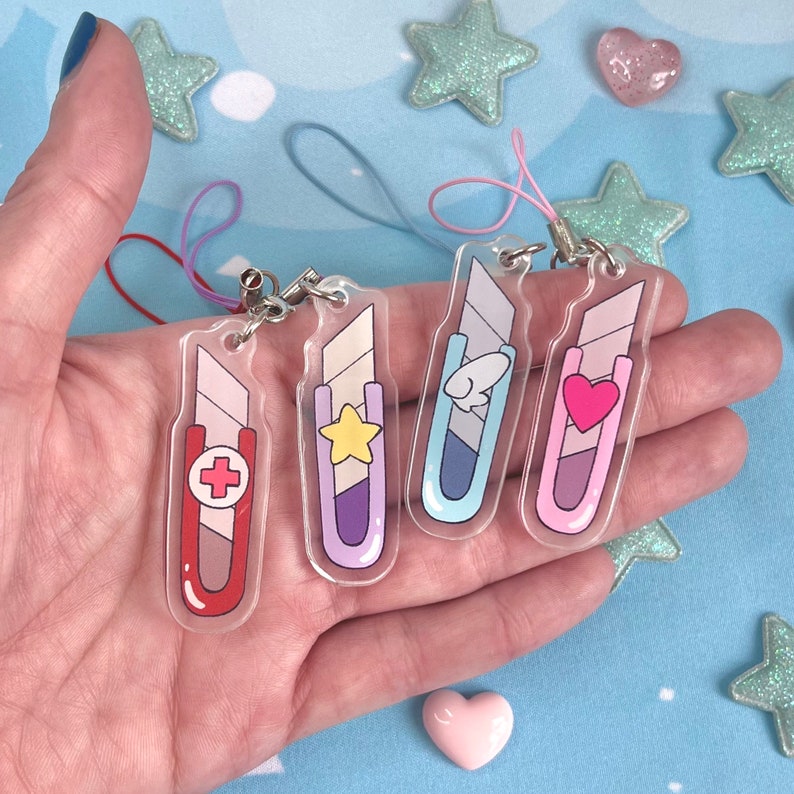 May include: Four clear acrylic charms shaped like utility knives, each with a different design: a red cross, a yellow star, a fish, and a pink heart. Each charm has a silver clasp and a colored string.