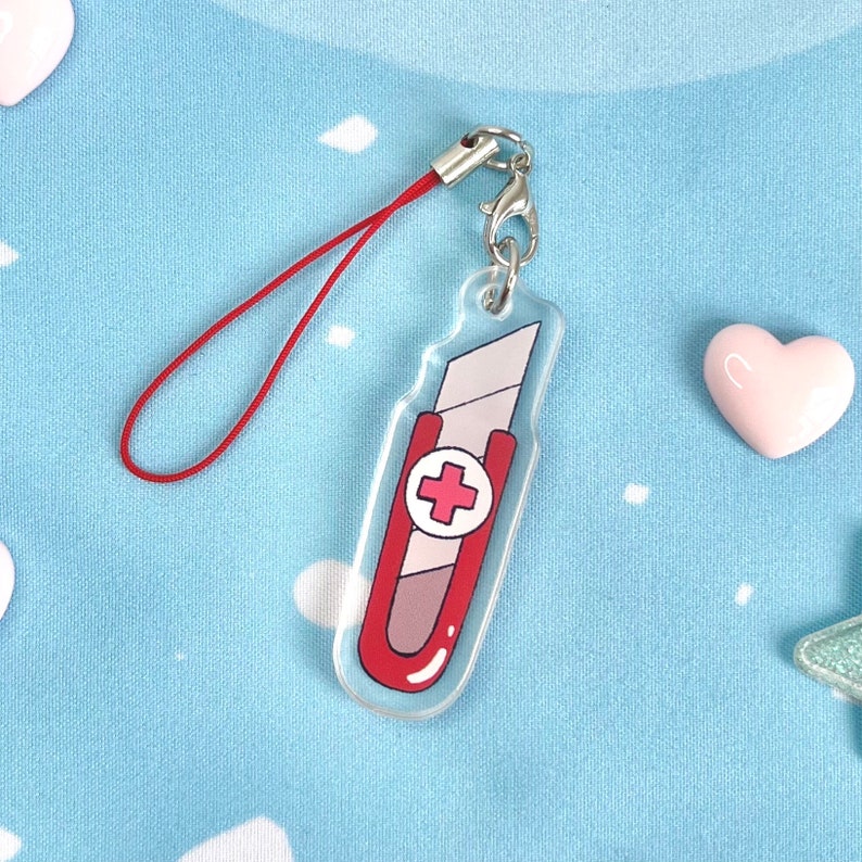 May include: A clear acrylic charm shaped like a red and gray utility knife with a red cross symbol. The charm has a silver clasp and a red lanyard. The background is a light blue fabric with white cloud shapes and pink heart-shaped objects.