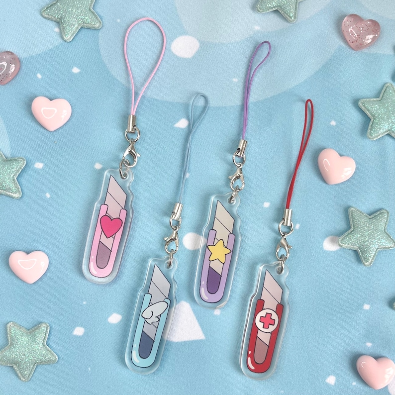 May include: Four acrylic charm keychains shaped like utility knives, each with a different design: a heart, a star, a cartoon character, and a medical cross. They have colored lanyards and silver clasps, set against a blue background with star and heart decorations.