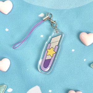May include: A clear acrylic charm in the shape of a utility knife, featuring a purple handle and a yellow star. It has a silver clasp and a purple lanyard. The charm is on a light blue background with heart-shaped decorations.