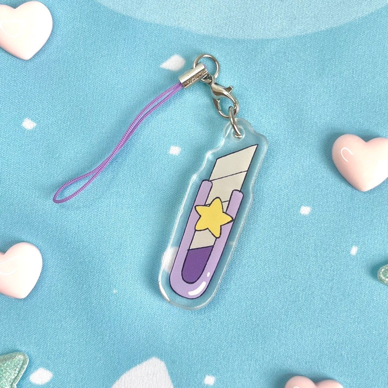 May include: A clear acrylic charm in the shape of a utility knife, featuring a purple handle and a yellow star. It has a silver clasp and a purple lanyard. The charm is on a light blue background with heart-shaped decorations.