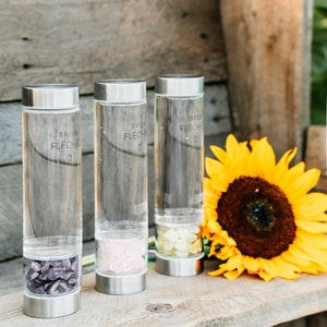 Könnte beinhalten: Drei Glaswasserflaschen mit Edelstahldeckeln und -böden. Jede Flasche enthält klares Wasser und eine Schicht bunter Kristalle am Boden. Die Flaschen sind mit "SOELA FLECHE" beschriftet und auf einer Holzoberfläche mit einer Sonnenblume im Vordergrund angeordnet.