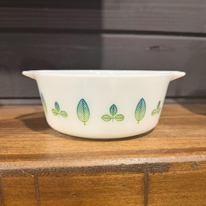 May include: White bowl featuring a leaf pattern in green and blue hues. The bowl has a rounded form with small handles. The leaf design is evenly spaced around the exterior, offering a decorative and practical kitchenware piece, perfect for serving.