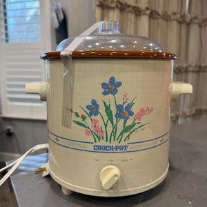 May include: Cream-colored Rival Crock-Pot slow cooker with a floral design in blue and pink. It has a brown rim and a clear glass lid. The control knob has settings for off, low, and high.