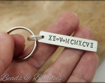 May include: Silver-colored rectangular keychain with a ring, featuring engraved Roman numerals and a heart symbol. The text reads XII♡V♡MCXCVII. The keychain is held in a hand, with the brand name "Beads & Beauties" visible at the bottom.
