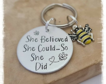May include: Silver keychain with a round charm that reads "She Believed She Could... So She Did" with a small bee design. A separate bee charm is attached to the keychain.