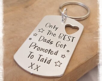 May include: A silver-colored metal keychain with a heart-shaped cutout and a ring. The keychain reads "Only The BEST Dads Get Promoted To Taid XX" with star accents. The words "Beads & Beauties" are at the bottom.