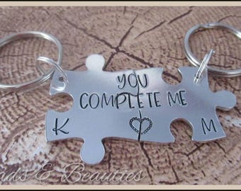 May include: Two silver puzzle pieces with key rings. The pieces fit together to form a heart and the text "You Complete Me". The left piece has the letter "K" engraved on it and the right piece has the letter "M" engraved on it.