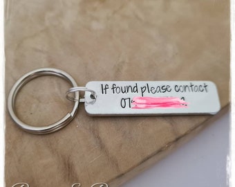 May include: Silver keychain with a rectangular tag that reads "If found please contact 07..." with a pink bar covering part of the phone number. The keychain has a silver ring.