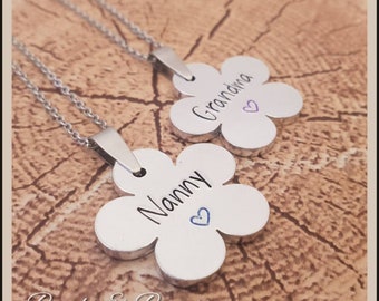 May include: Two flower-shaped pendants with the words "Nanny" and "Grandma" engraved in black script. Each pendant has a small blue heart and hangs from a silver chain. The background is a wood grain pattern.