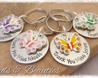 May include: Keychains with butterfly charms in various colors, including purple, pink, yellow, and green. Each keychain has a round silver tag with the words "Thank You Miss" and a name. The text "Beads & Beauties" is also visible.