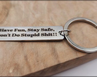 May include: A silver-colored keychain with a rectangular tag and a circular key ring. The tag is engraved with the phrase "Have Fun, Stay Safe, Don't Do Stupid Shit!!" in a bold, sans-serif font. The keychain rests on a wooden surface.