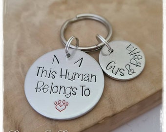 May include: Silver keychain with two round tags. The larger tag says "This Human Belongs To" with a red heart and paw print. The smaller tag says "Gus & Bella".