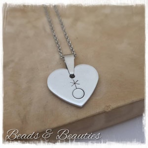 May include: A silver heart-shaped pendant necklace with a symbol of a circle with a cross on top. The necklace is on a silver chain.