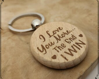 May include: A wooden keychain with a silver ring. The round wooden pendant has the engraved text "I Love You More... The End I WIN" with heart symbols. The keychain is light brown.