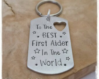 May include: Silver keychain with a heart cutout and the text "To The ☆ BEST ☆ First Aider ☆ In The ☆ World ☆".