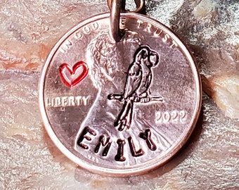 Op de afbeelding: Een koperen sleutelhanger met een klein rood hartje, een vogelontwerp en de naam "Emily" erop gegraveerd. Het jaar 2022 is ook gegraveerd op de cent. De sleutelhanger is bevestigd aan een zilveren sleutelring.