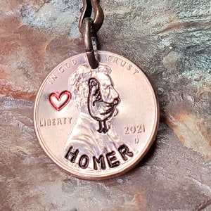 May include: A copper penny keychain with a red heart and the word "HOMER" engraved on it. The penny is from 2021 and features the profile of Abraham Lincoln.