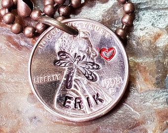 Op de afbeelding: Een koperen penny ketting met een libellenontwerp en de naam "Erik" erop gegraveerd. De ketting heeft een koperen balketting en een rode hartbedel.
