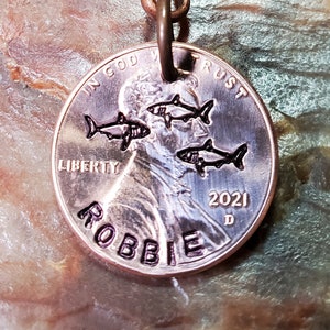 Op de afbeelding: Een koperen sleutelhanger met een gepersonaliseerde inscriptie "ROBBIE" en het jaar "2021". De penny toont een afbeelding van Abraham Lincoln en de woorden "In God We Trust" en "Liberty". Vier kleine vissen zijn gegraveerd op de penny.