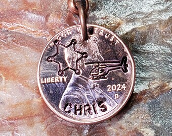 Op de afbeelding: Een koperkleurige sleutelhanger met een 2024 penny waarop een helikopter en een sheriffster staan afgebeeld. De penny heeft de woorden "In God We Trust" en "Liberty" erop. De sleutelhanger heeft ook de naam "CHRIS" gegraveerd op de penny.