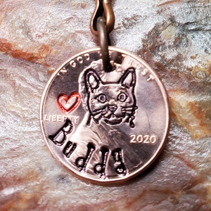 May include: A copper penny keychain with a cat's face engraved on it. The cat's eyes are black and the penny has the words "In God We Trust" and "Liberty" engraved on it. The year 2020 is also engraved on the penny. The keychain has a copper ring and a copper jump ring. The word "Buddy" is engraved on the penny.