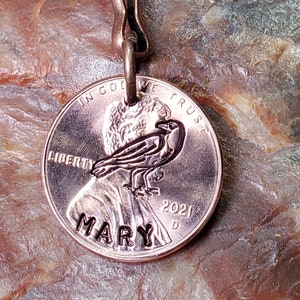 May include: A copper penny keychain with a personalized inscription of "MARY" and the year "2021". The penny features a design of a bird with the words "IN GOD WE TRUST" and "LIBERTY" on the coin.