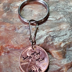 Op de afbeelding: Een koperen sleutelhanger met een zilveren sleutelring. De penny is gegraveerd met het woord "JOSH" en een ontwerp van vijf vissen. De penny is gedateerd 1983 en heeft de woorden "IN GOD WE TRUST" en "LIBERTY" erop gegraveerd.